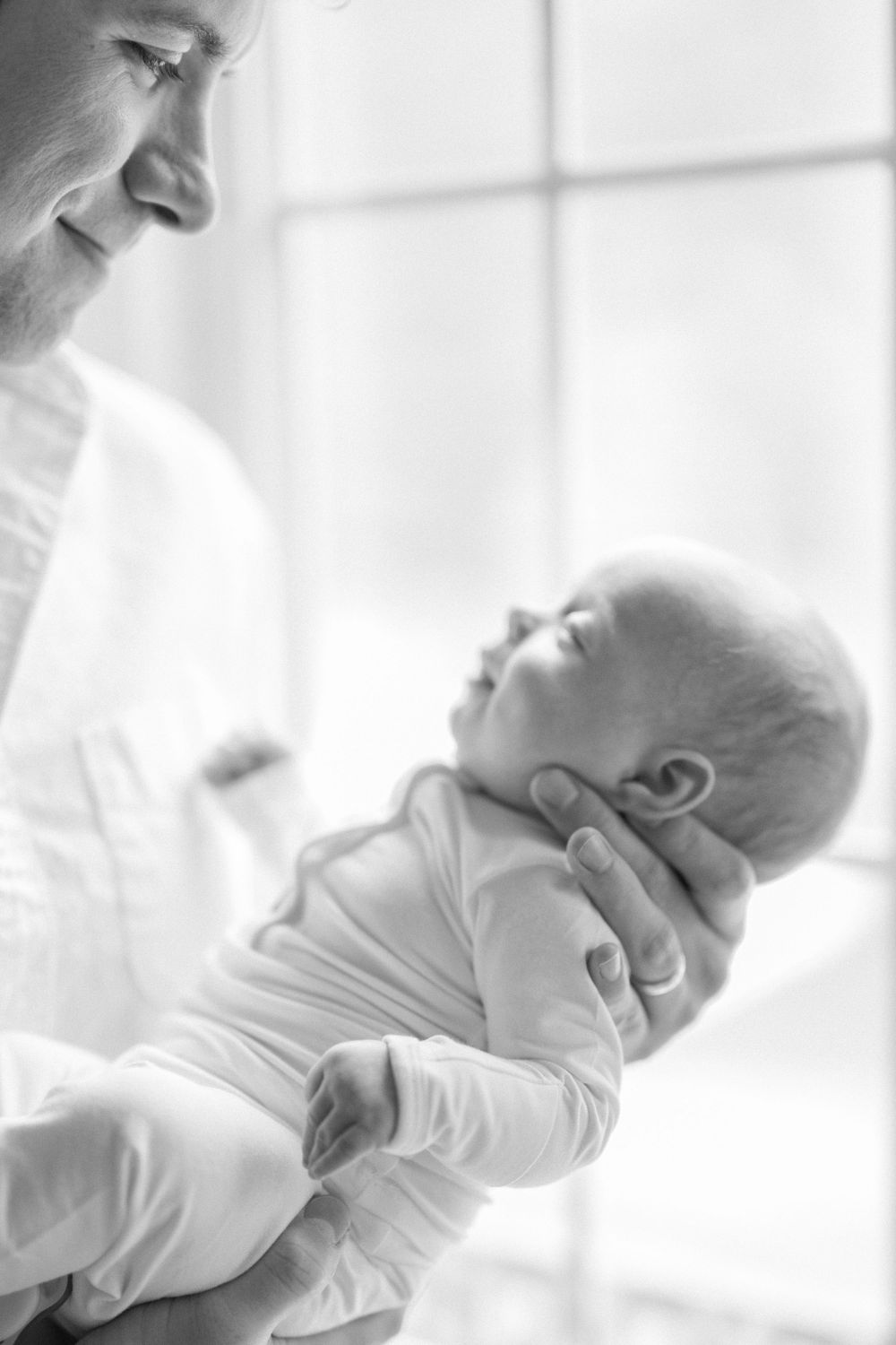 A black and white photo of a parent cradling a newborn baby in white clothing near a window taken by a northern virginia newborn photographe
