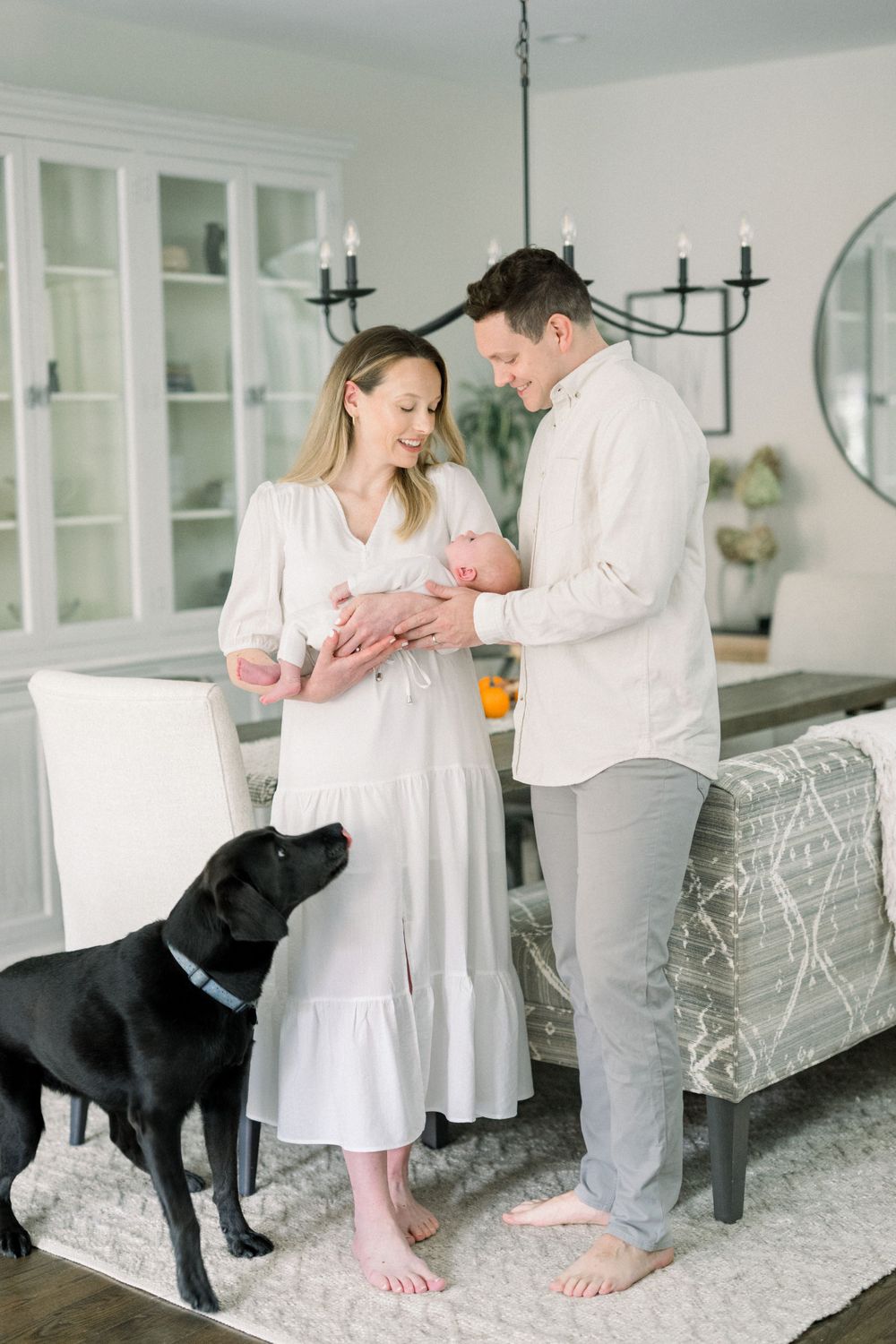 A couple in white clothing stands with their baby in a bright living room while their black Labrador dog looks on.