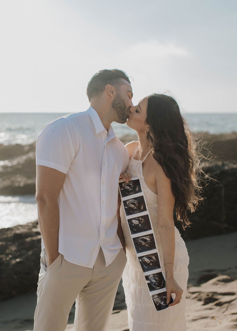 Romantic beach pregnancy announcement photoshoot at sunset with couple holding ultrasound photos.