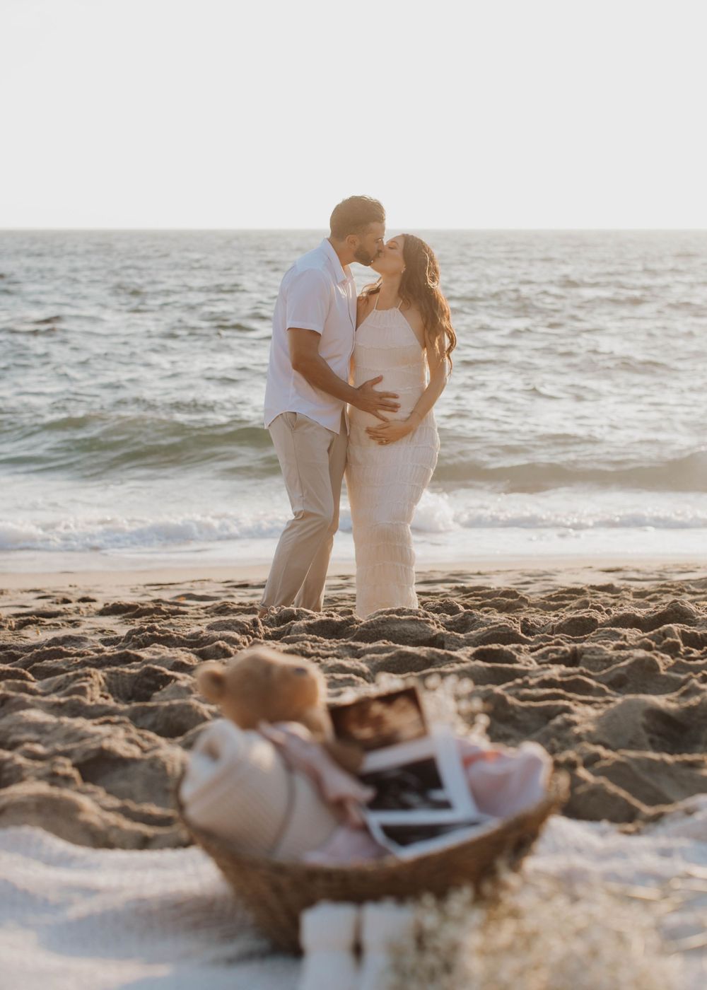 Couple celebrating on sandy beach with ultrasound photos and romantic sunset backdrop.