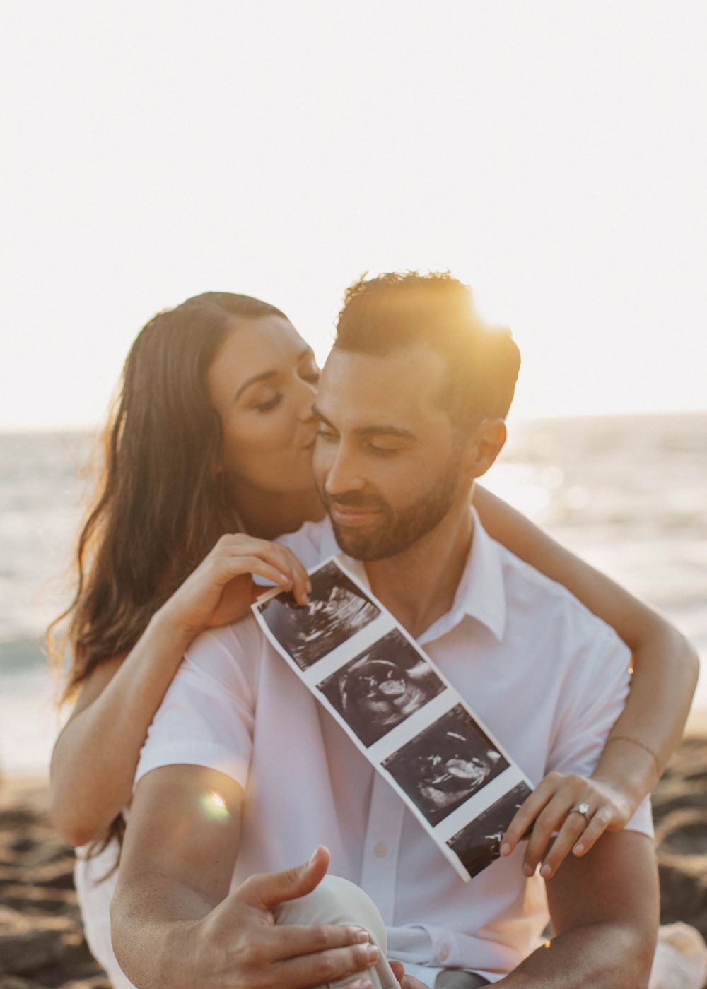 Sunset beach photo session with couple sharing tender moments while holding ultrasound images.