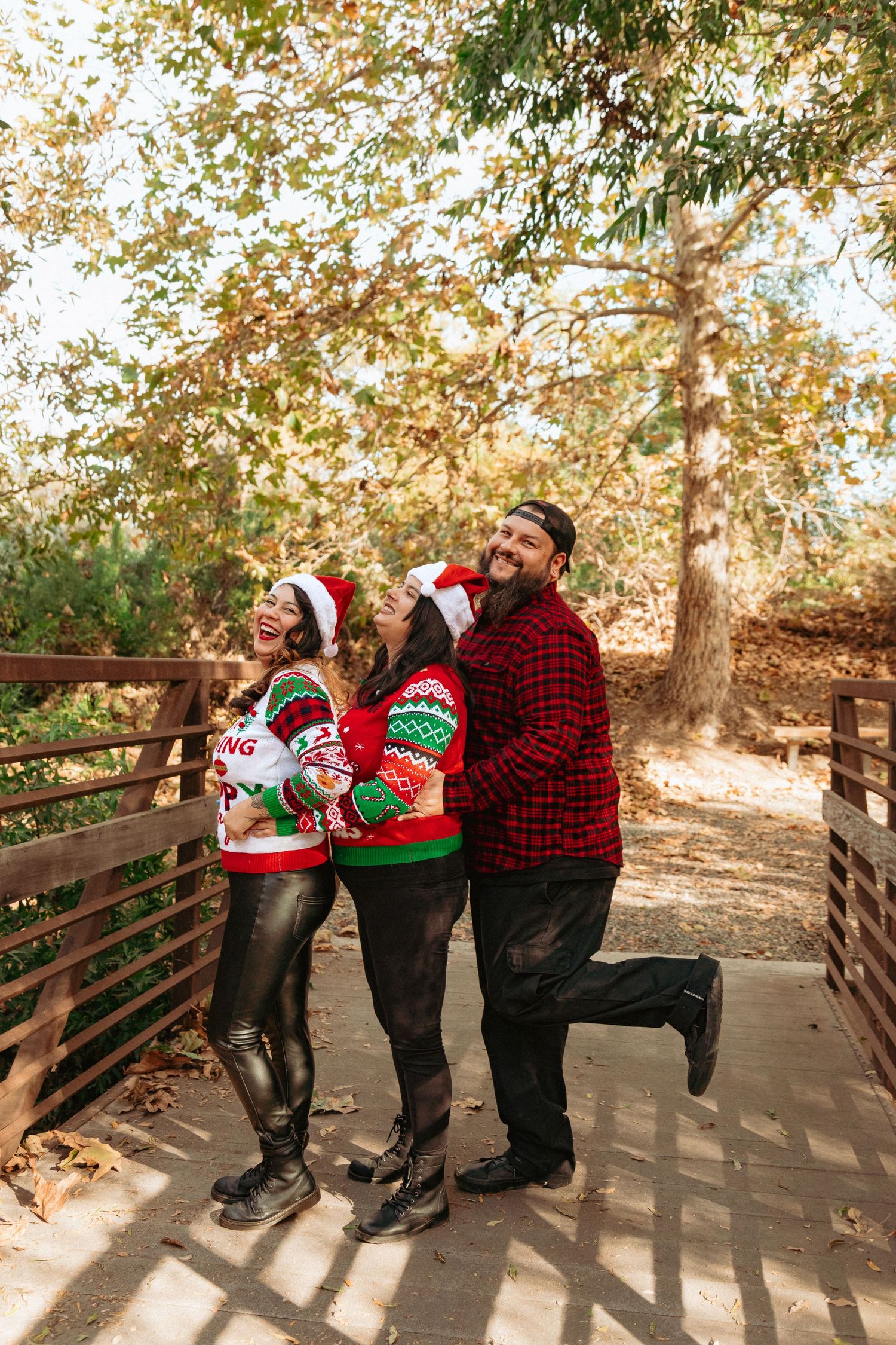 Siblings in festive Christmas sweaters and Santa hats stand laughing together on a wooden bridge during autumn.