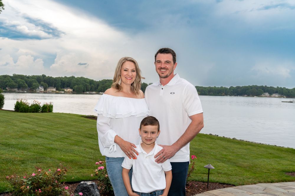 Family posing outdoors near a scenic lake with lush green lawn and dramatic cloudy sky in the background.