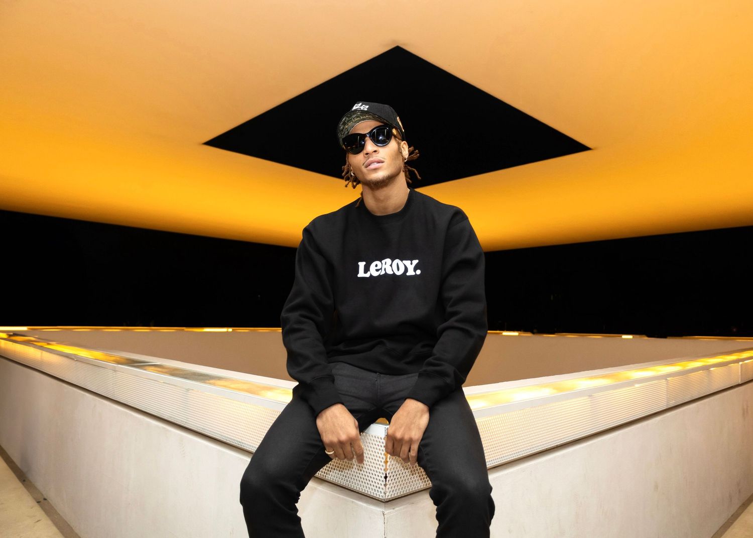 Person wearing black LXROY hoodie sits beneath black geometric ceiling against orange backdrop.