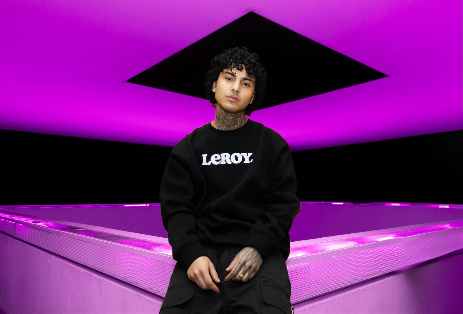 Person in black LXROY hoodie sits under purple lighting with geometric black ceiling feature.