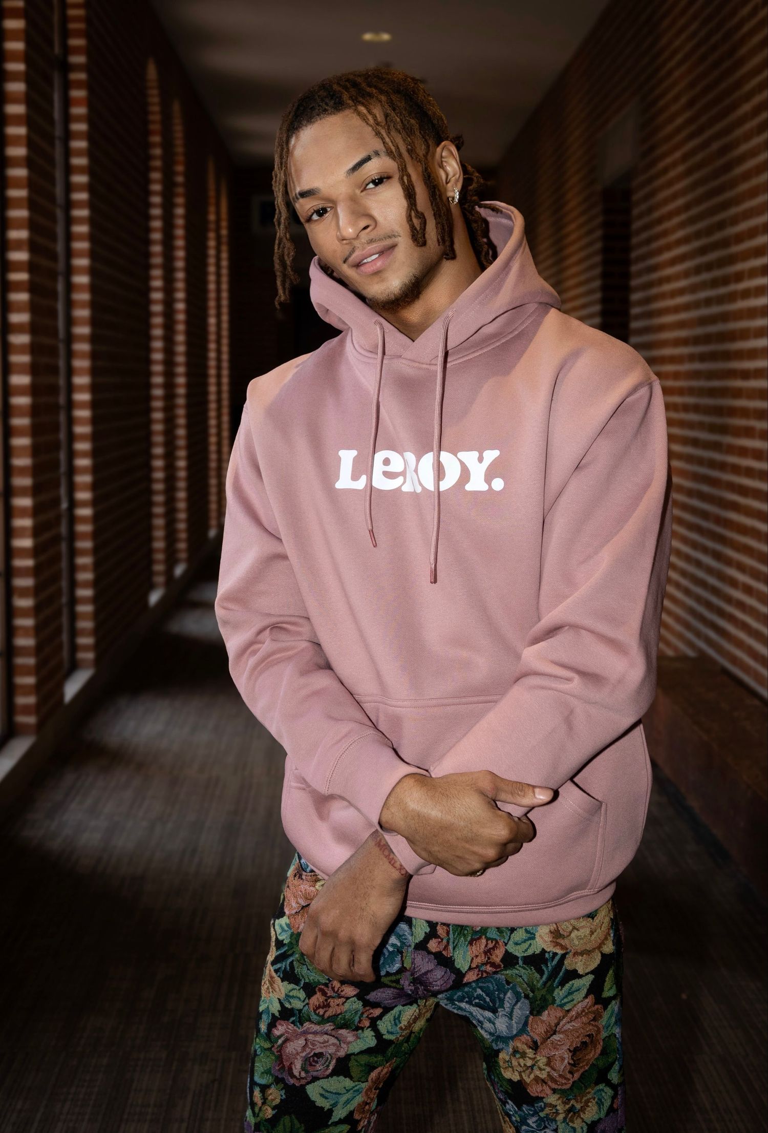 Person wearing pink LXROY hoodie and floral pants poses in dimly lit hallway.