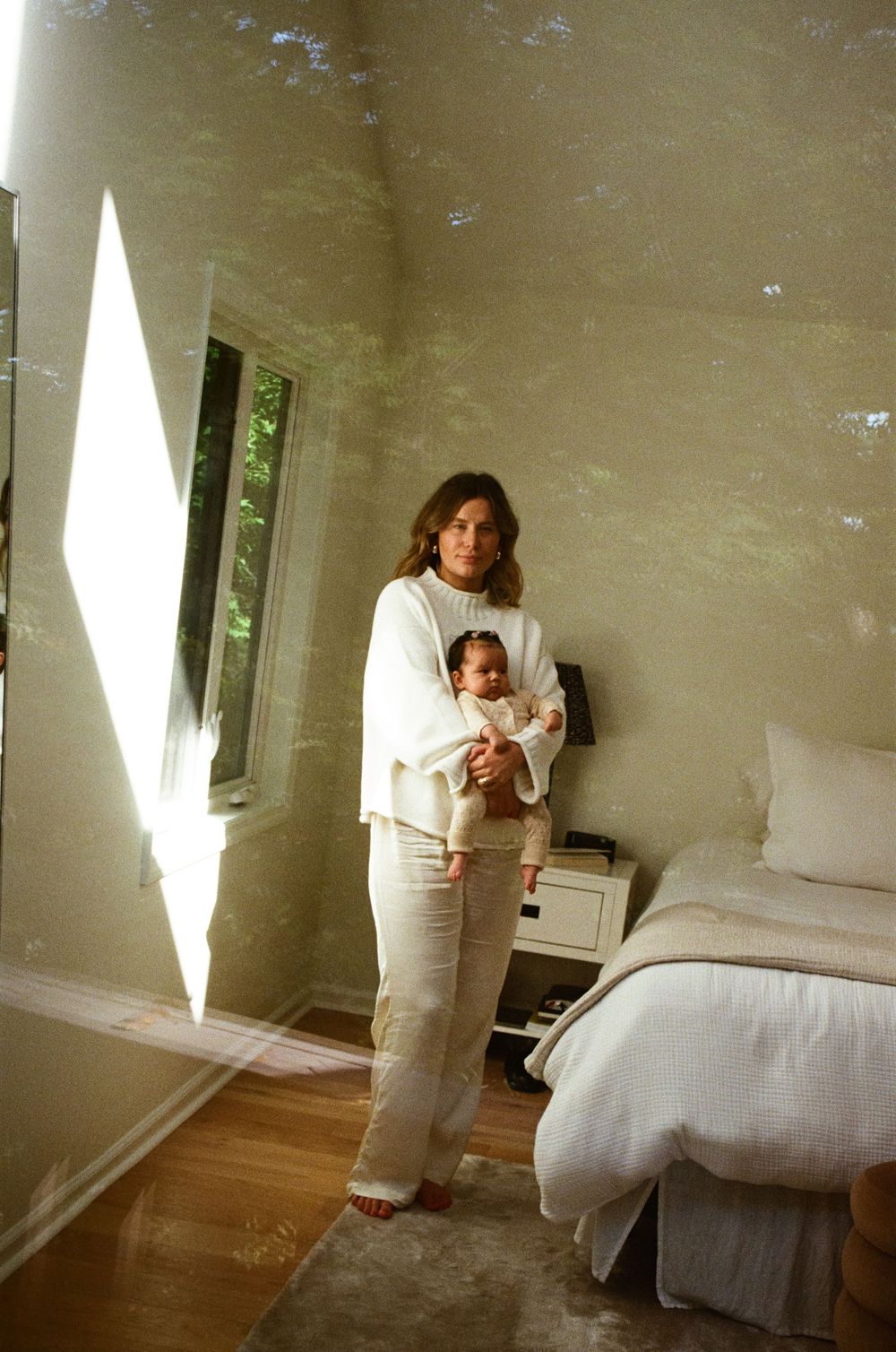 Woman in white outfit holds newborn baby in sunlit bedroom with hardwood floors and neutral decor.