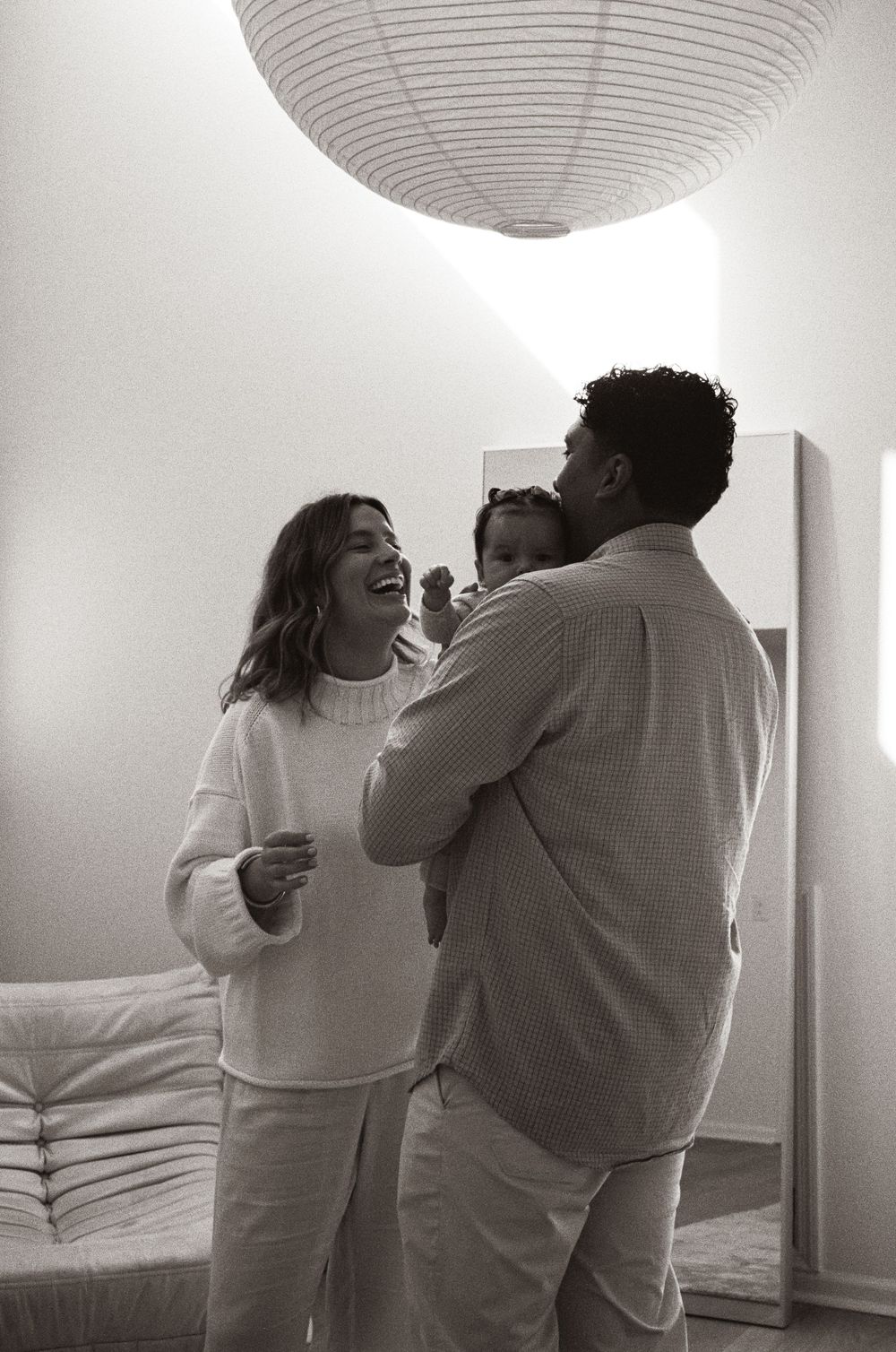 Black and white photo of a happy couple holding a baby, laughing together in a bright bedroom.