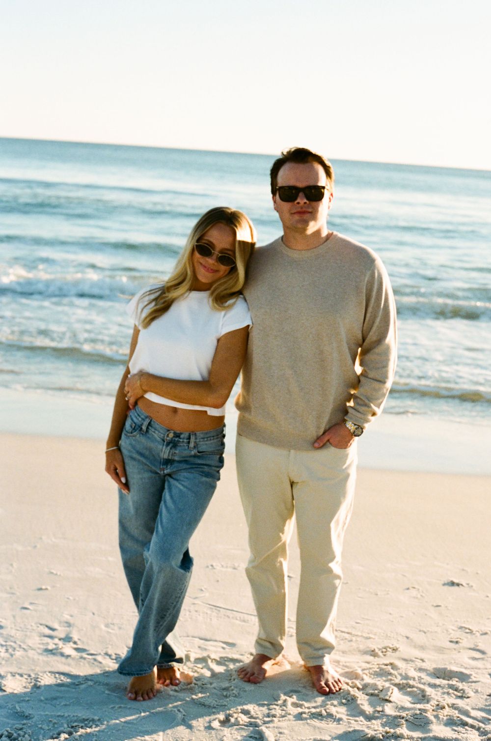 A couple wearing sunglasses poses barefoot on a sandy beach, woman in white crop top and jeans, man in beige sweater.