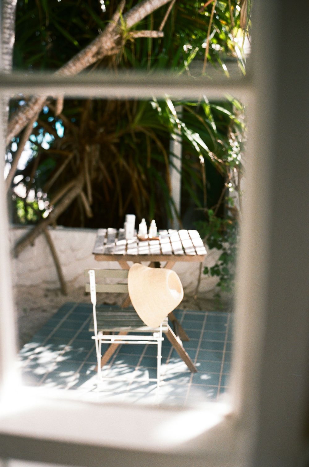 Outdoor patio table with bottles viewed through window, surrounded by tropical trees on blue tile floor.