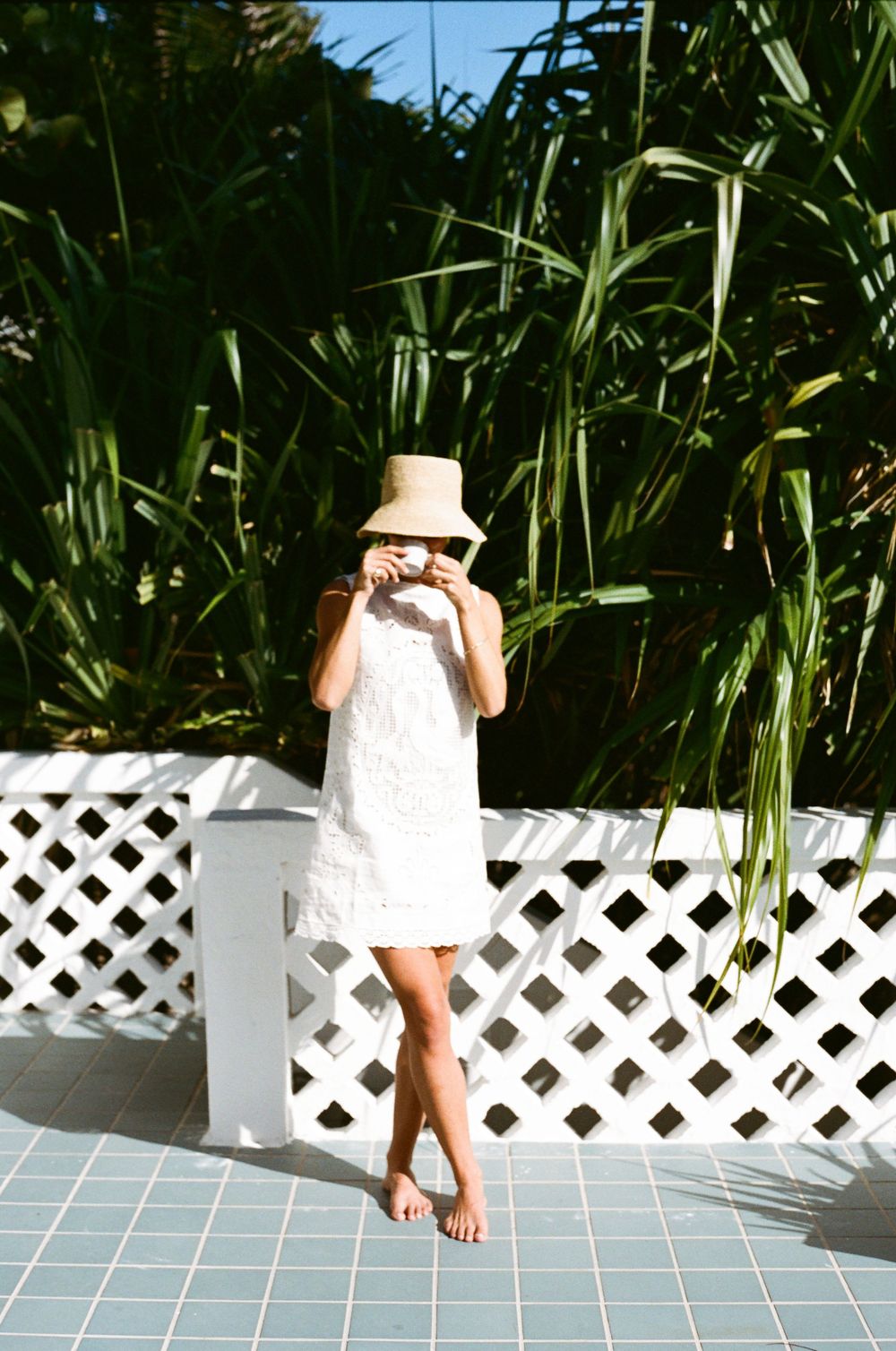 Woman in white dress and straw hat standing barefoot on blue tiles by white lattice fence with tropical greenery.
