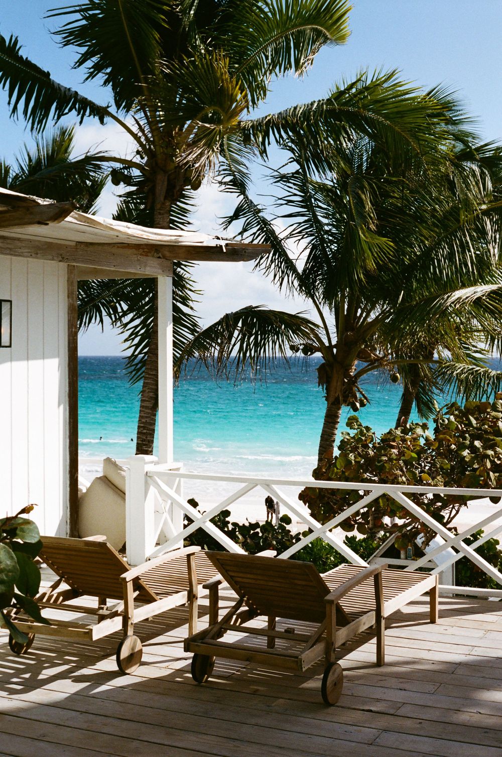 Two wooden lounge chairs on a tropical beachfront deck with palm trees and turquoise ocean views.
