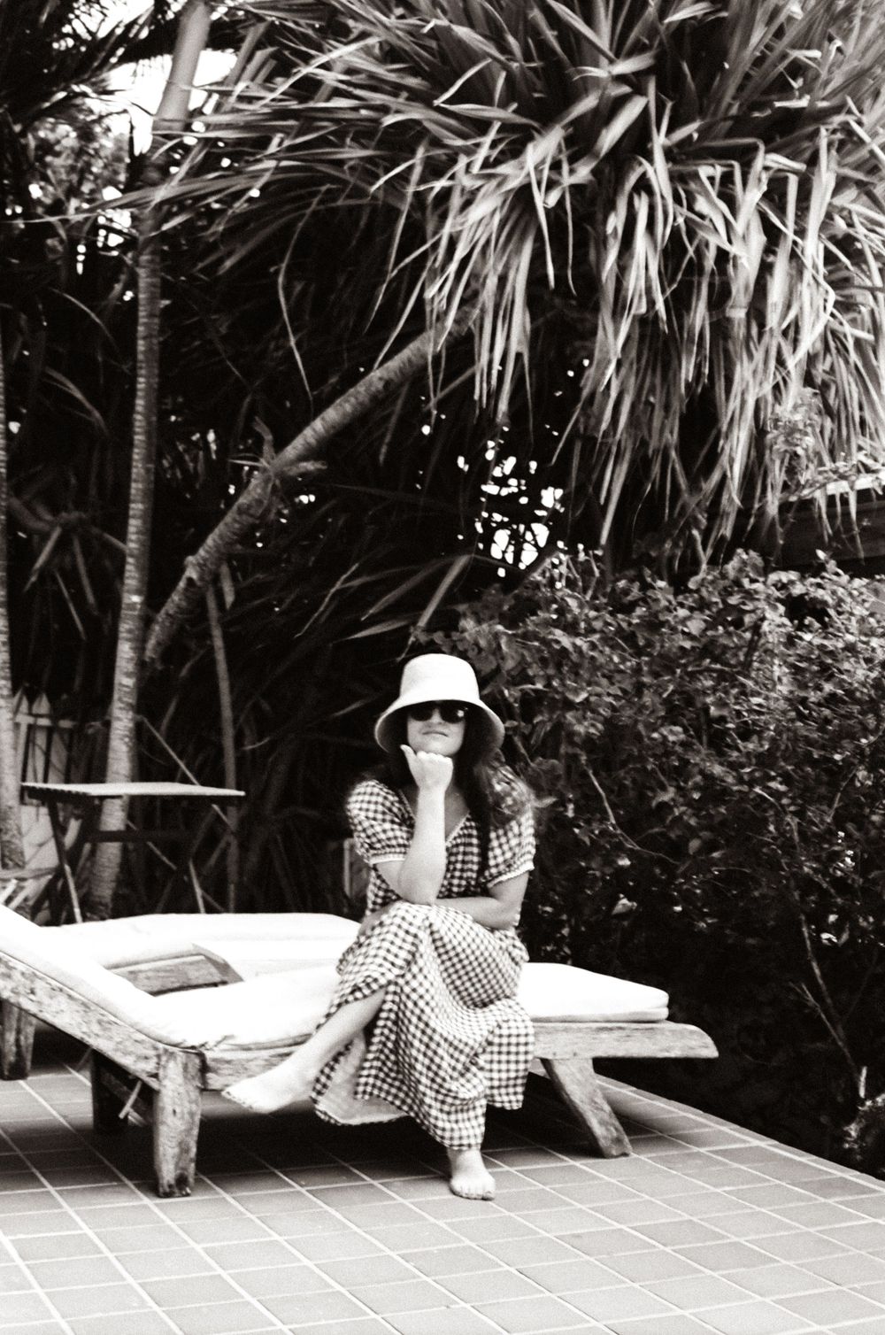 Woman in checkered dress and sun hat sits on wooden lounge chair surrounded by tropical palms in black and white photo.