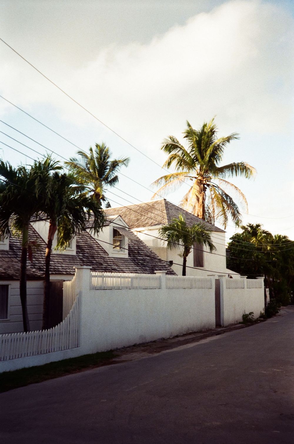 White picket fence beside a quiet street with palm trees and a classic tropical house under a partly cloudy sky.