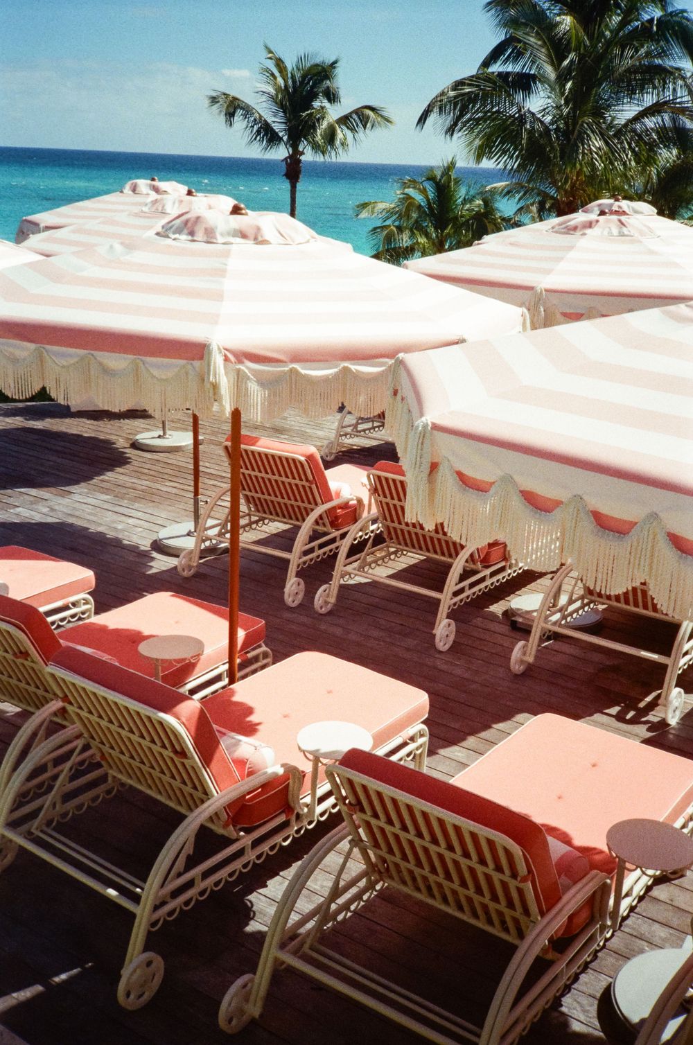 Pink striped beach umbrellas with white wicker lounge chairs on a wooden deck overlooking a tropical turquoise ocean.
