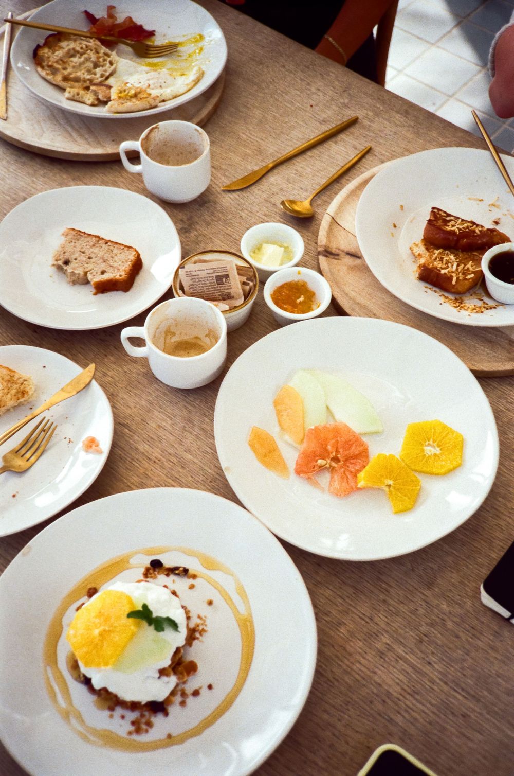 Overhead view of a brunch spread with citrus fruit plate, poached egg dish, coffee cups, toast, and condiments on a wooden table.