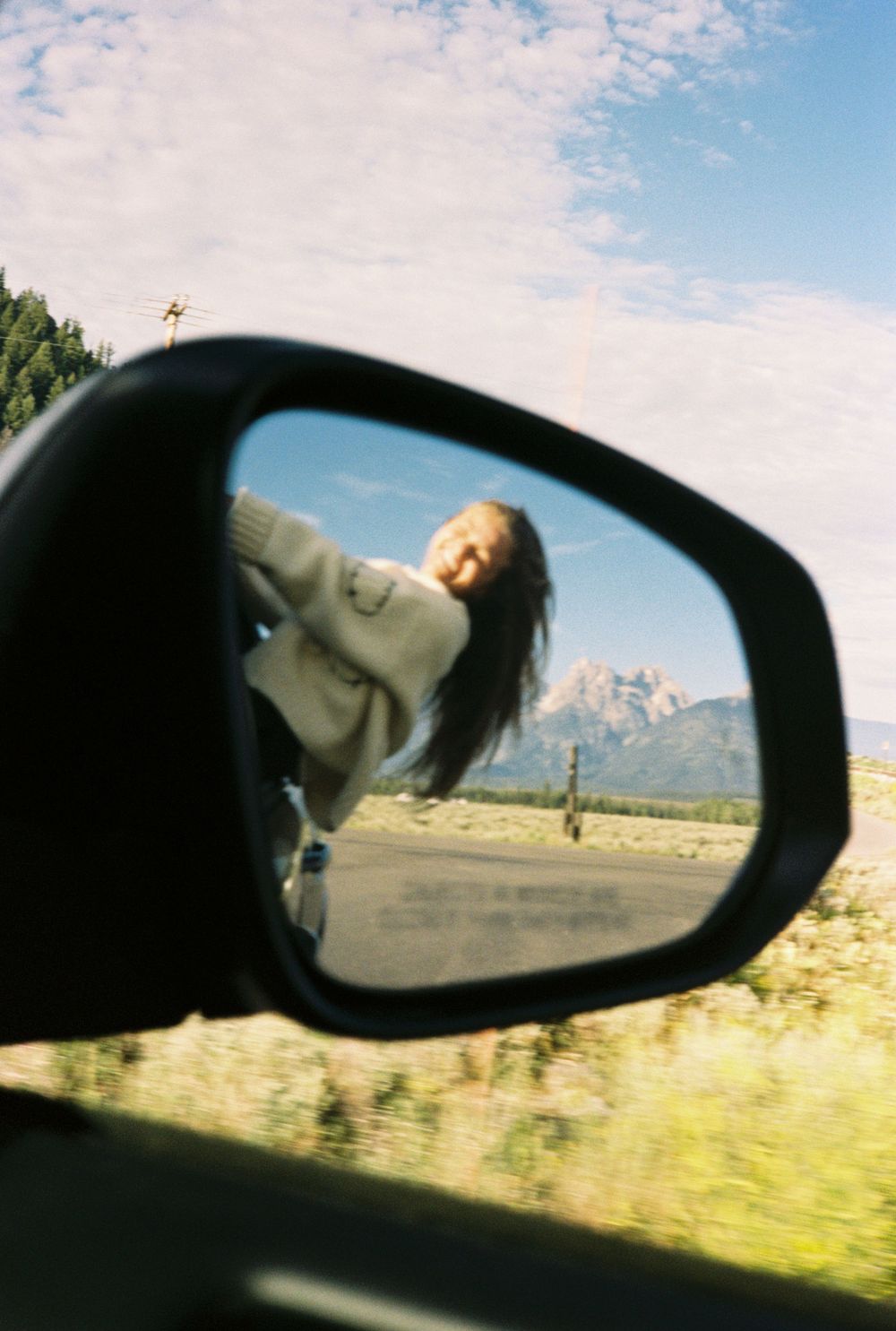 Woman leaning out car window reflected in side mirror with mountain landscape backdrop.