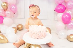 Pink and white balloons decorate a birthday cake smash photo session with a small child in a tutu dress.