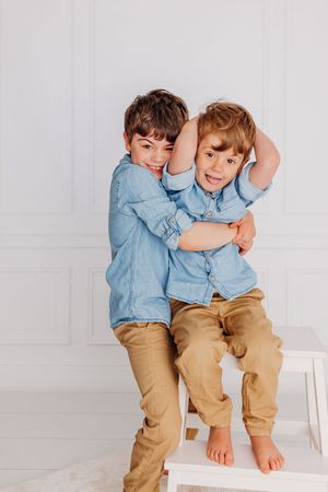 Two children in matching denim shirts and khaki pants sit playfully together on a white bench.