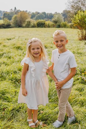 Two young children in white outfits pose together in a sunny meadow with tall grass and trees in the background.