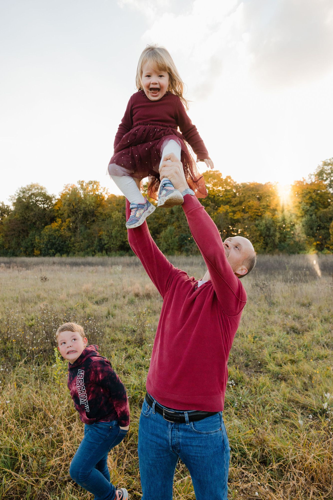 Family playing together in a sunlit meadow during golden hour, showing playful lifting movements.