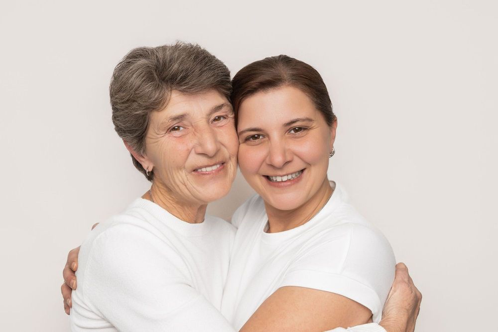 Two people wearing white shirts embrace warmly while smiling at the camera in a bright studio setting.