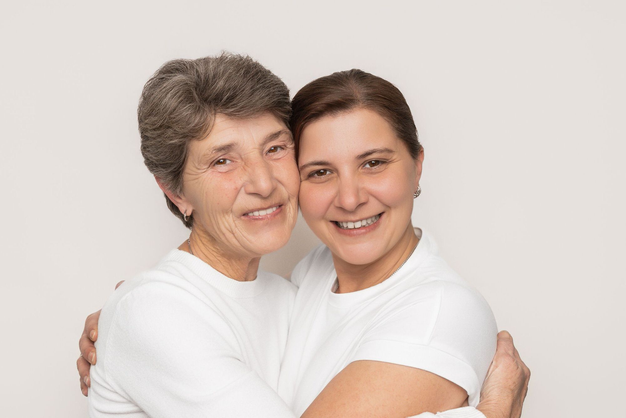 Two people wearing white shirts embrace warmly while smiling at the camera in a bright studio setting.