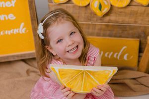 Child in pink dress holds decorative yellow lemon slice pillow against rustic wooden backdrop with pumpkins.
