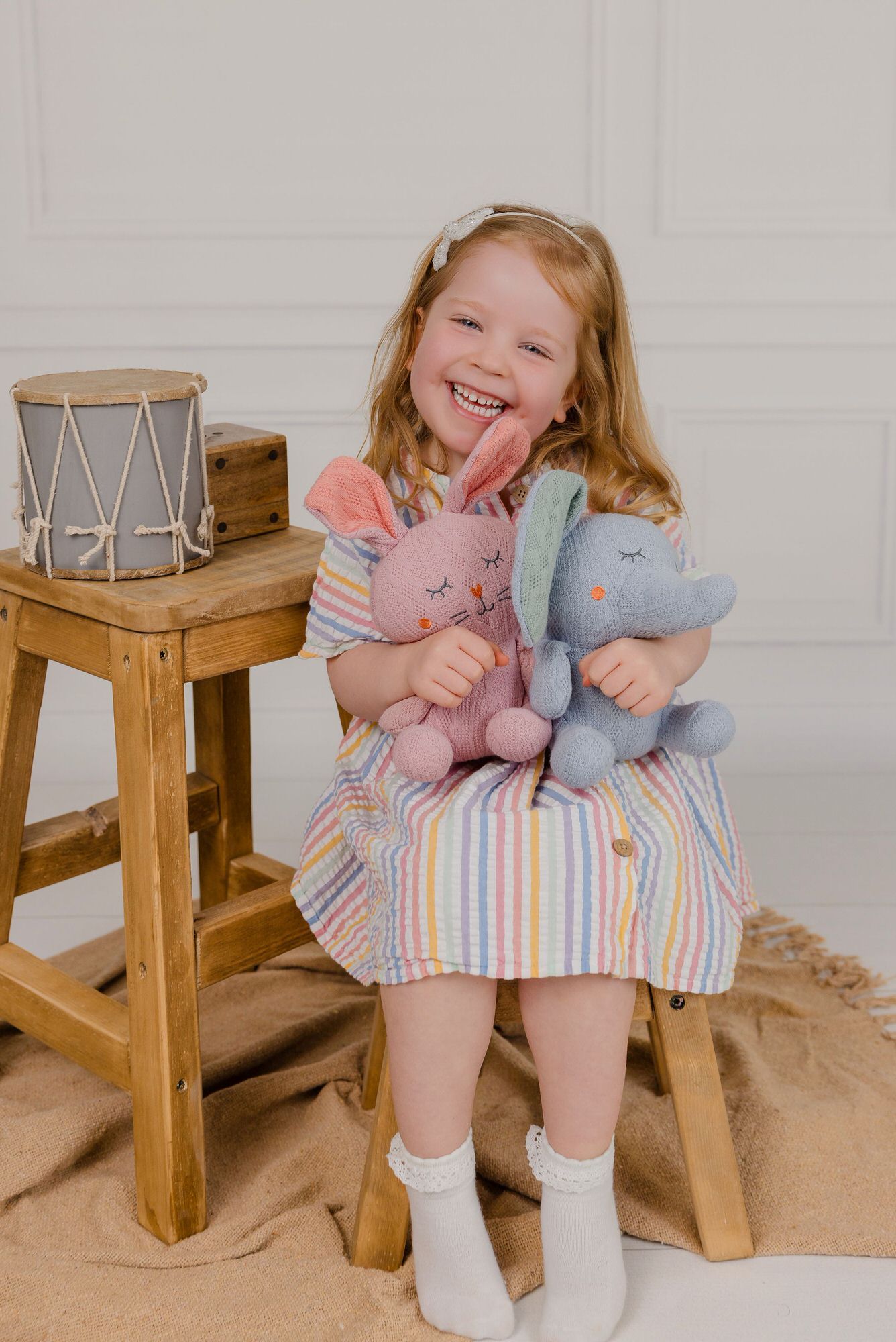 Small wooden stool setup with soft plush toys and rainbow striped decor against a white wall.