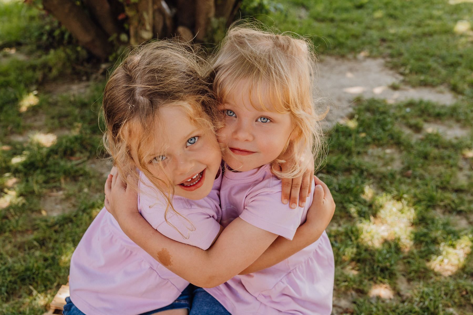 Two children in matching pink shirts share a tender hug while sitting outdoors in a garden setting.