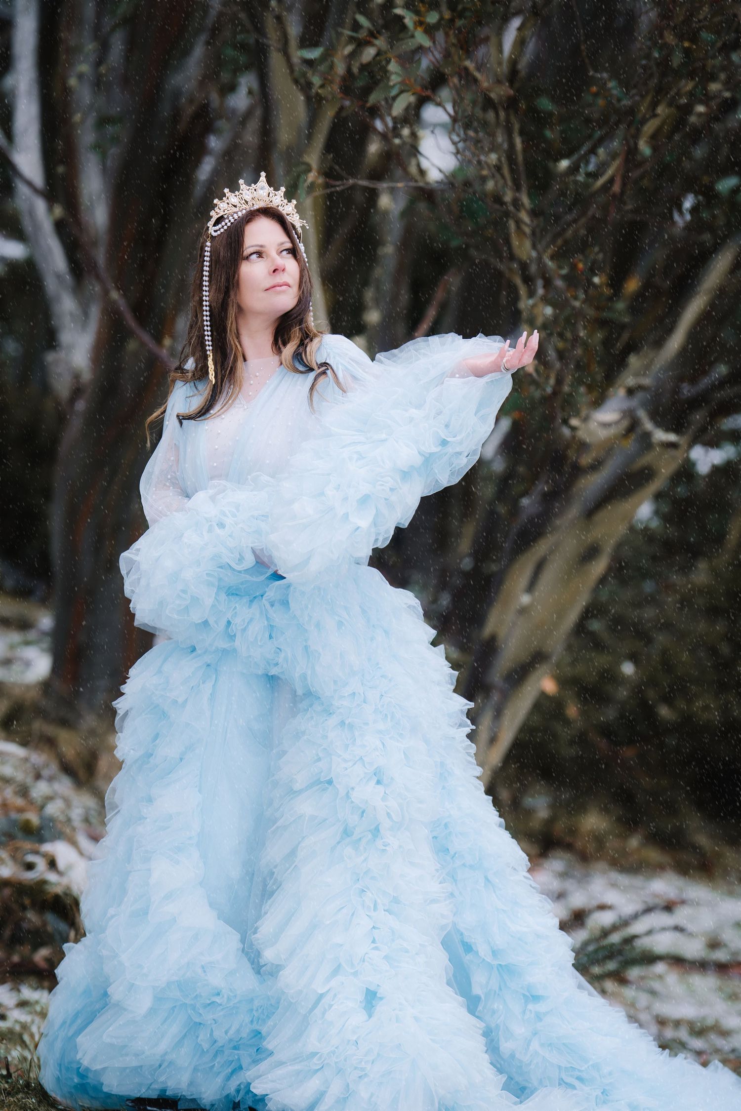 Woman in luxurious pale blue gown  and pearl headpiece poses amid trees while snow is falling around her.