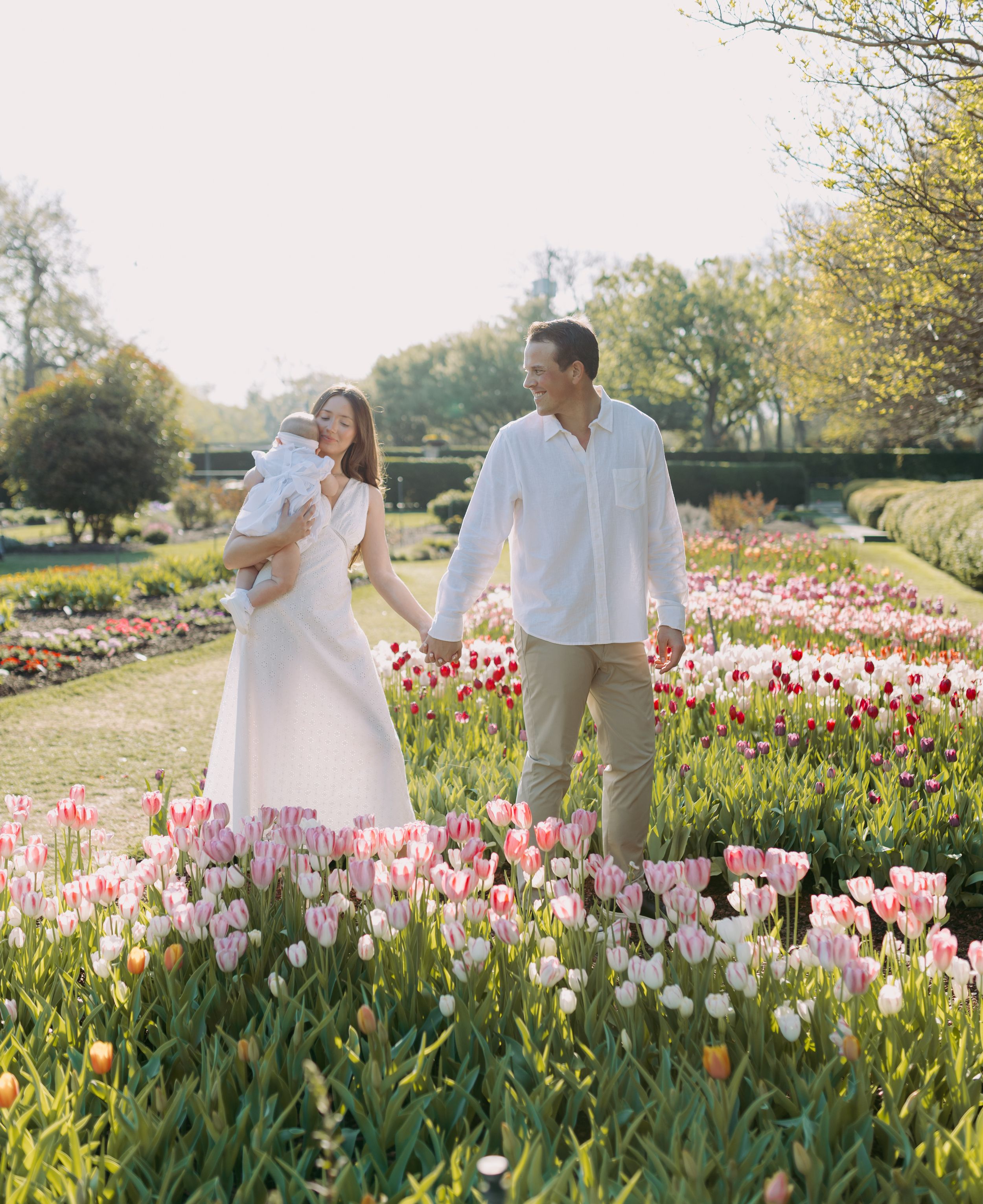 A couple in white attire strolls through a vibrant garden filled with blooming pink and yellow tulips on a sunny spring day.