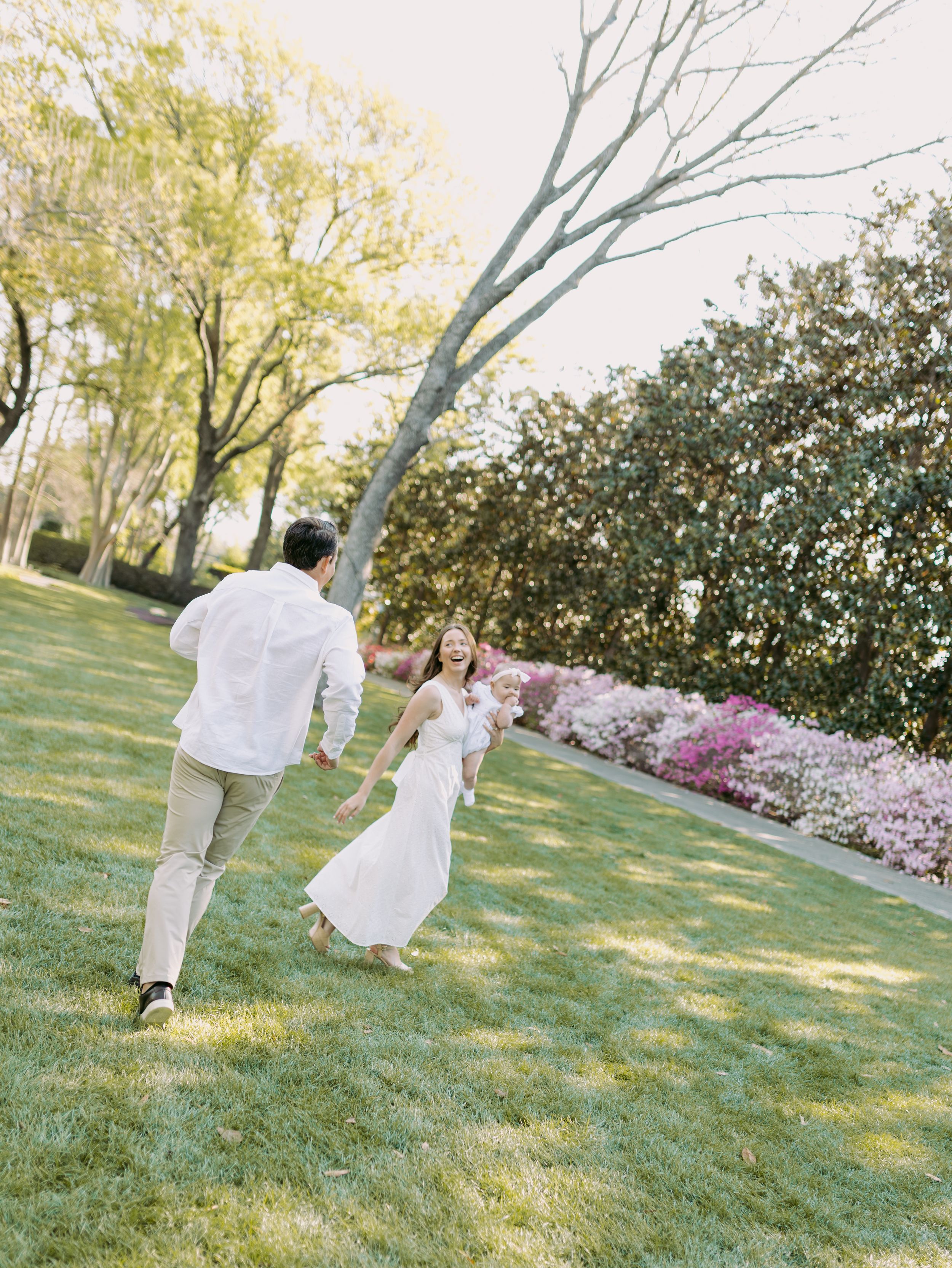 Couple in white outfits playfully dance and twirl on a grassy lawn with blooming trees in background.
