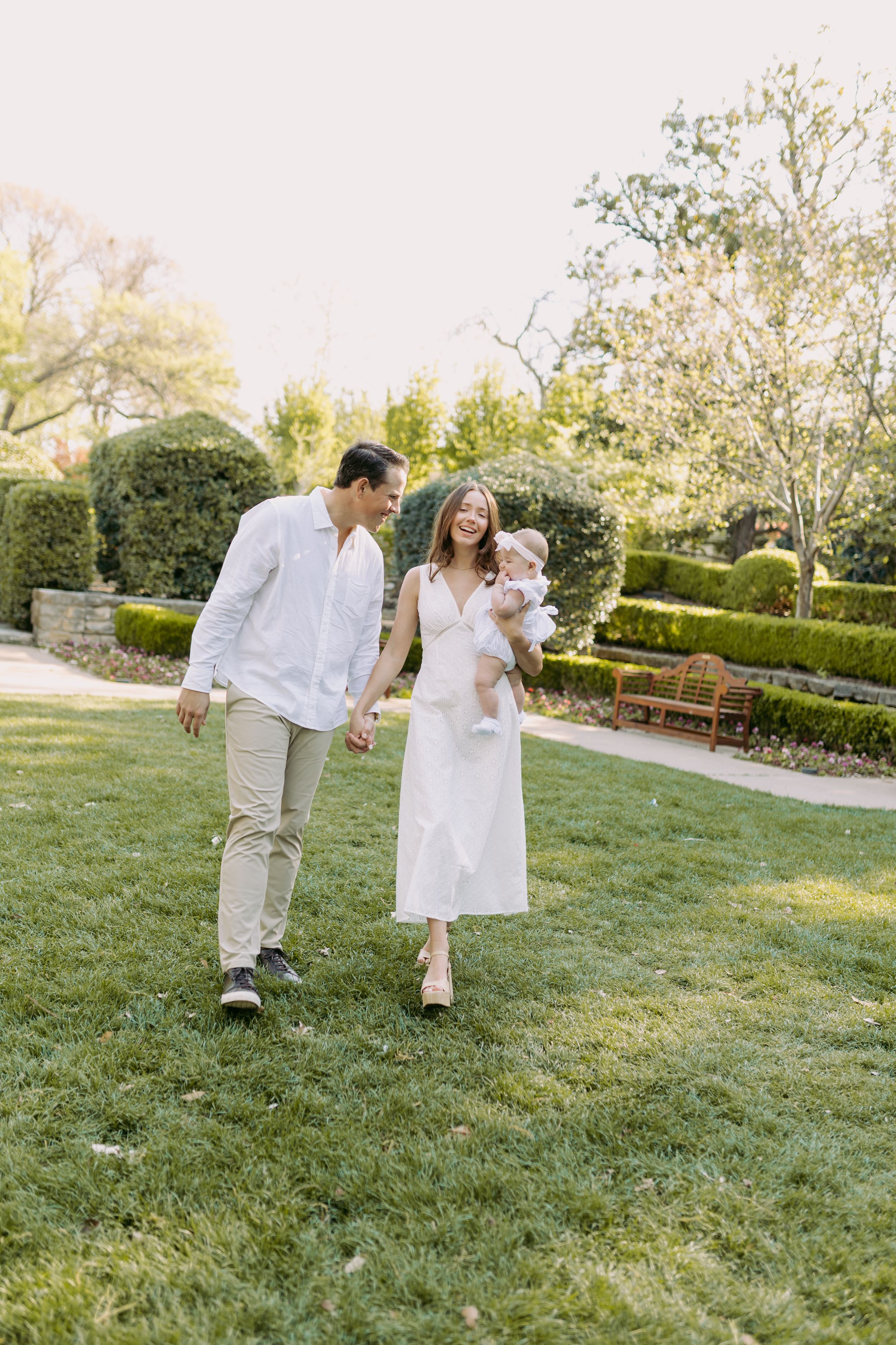 A family strolling together through a garden park, wearing coordinated white and beige outfits on a sunny day.