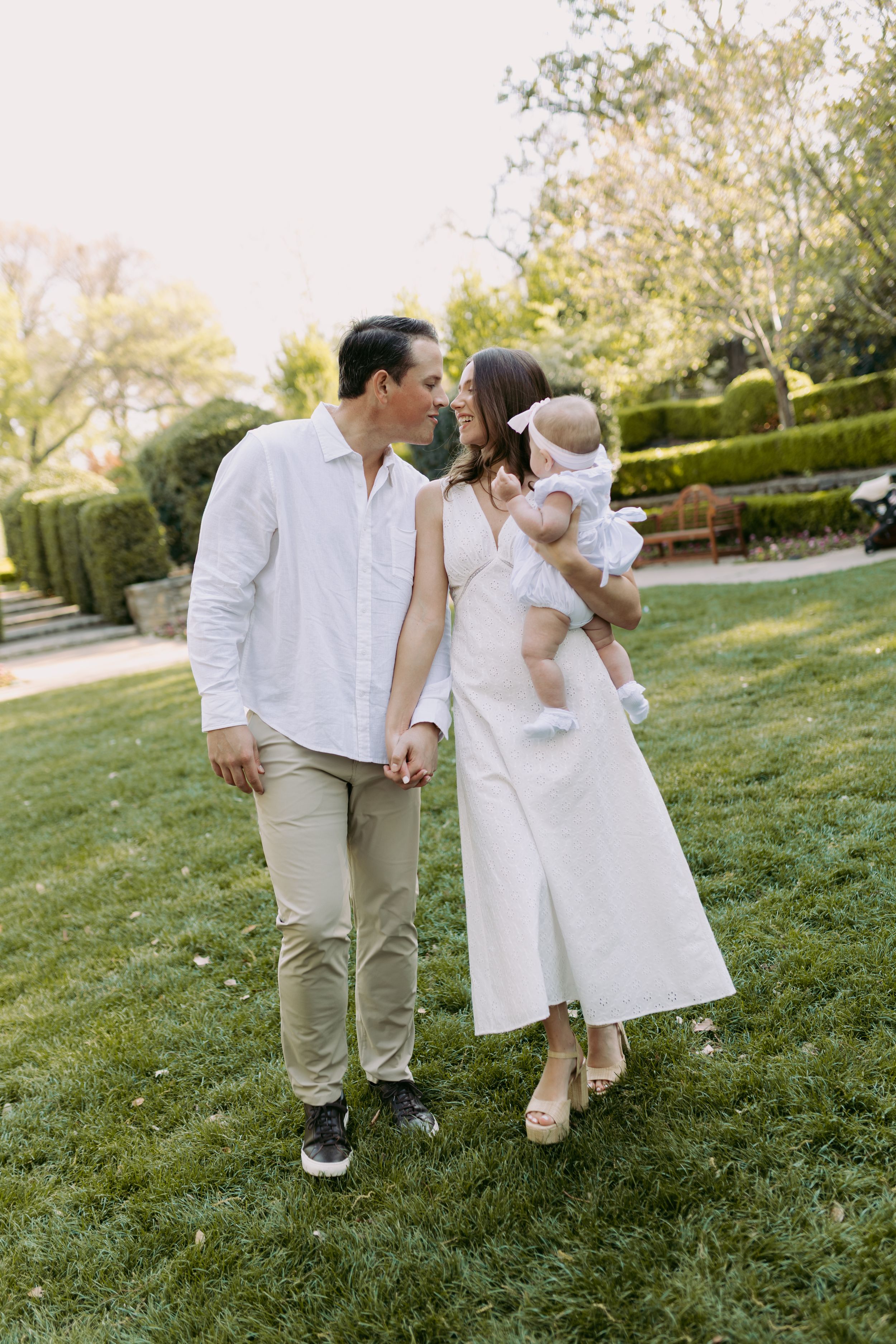 A couple in white outfits sharing a tender moment with their baby on a manicured lawn with hedges in the background.