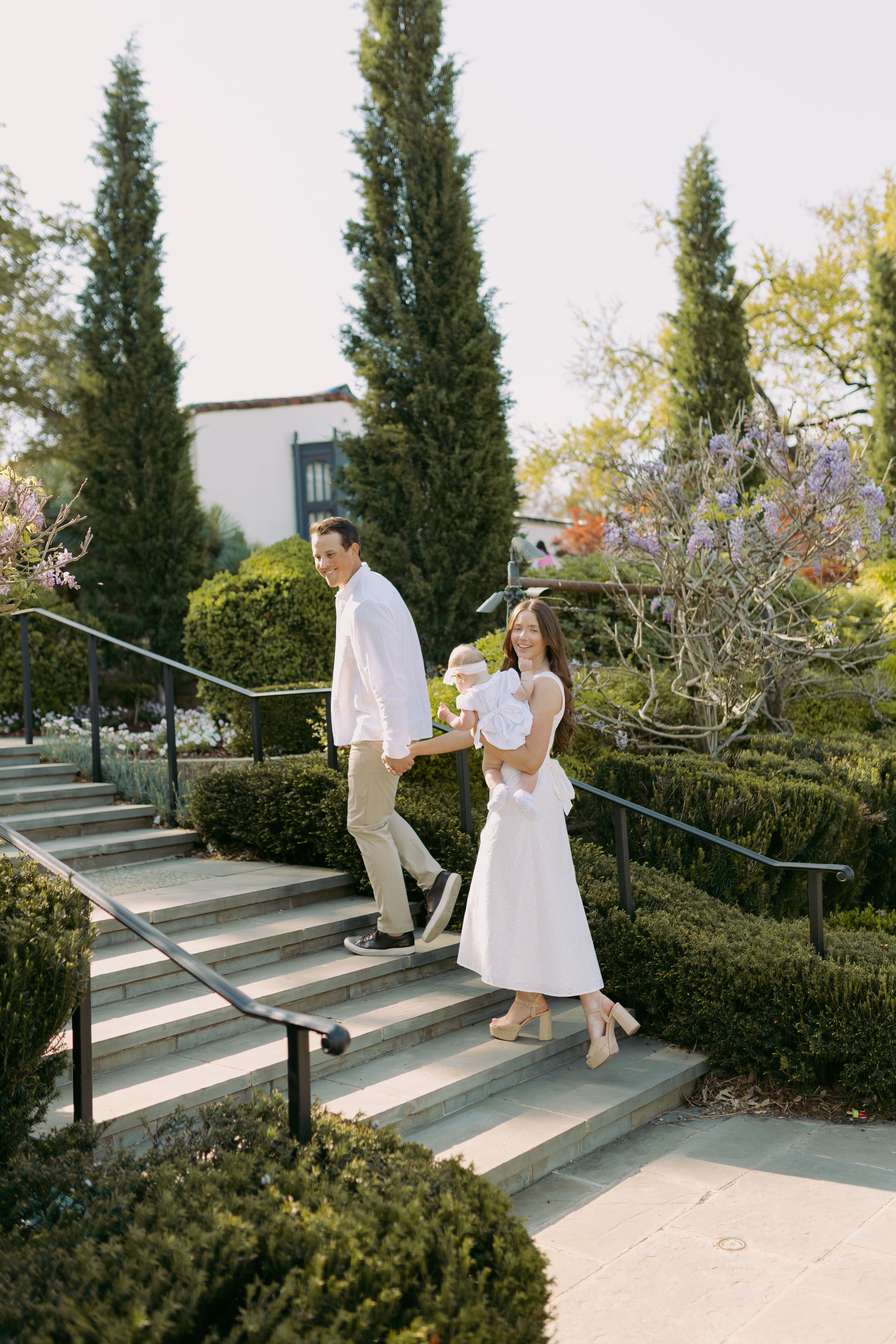 Couple in white attire descends concrete steps surrounded by manicured hedges and cypress trees.