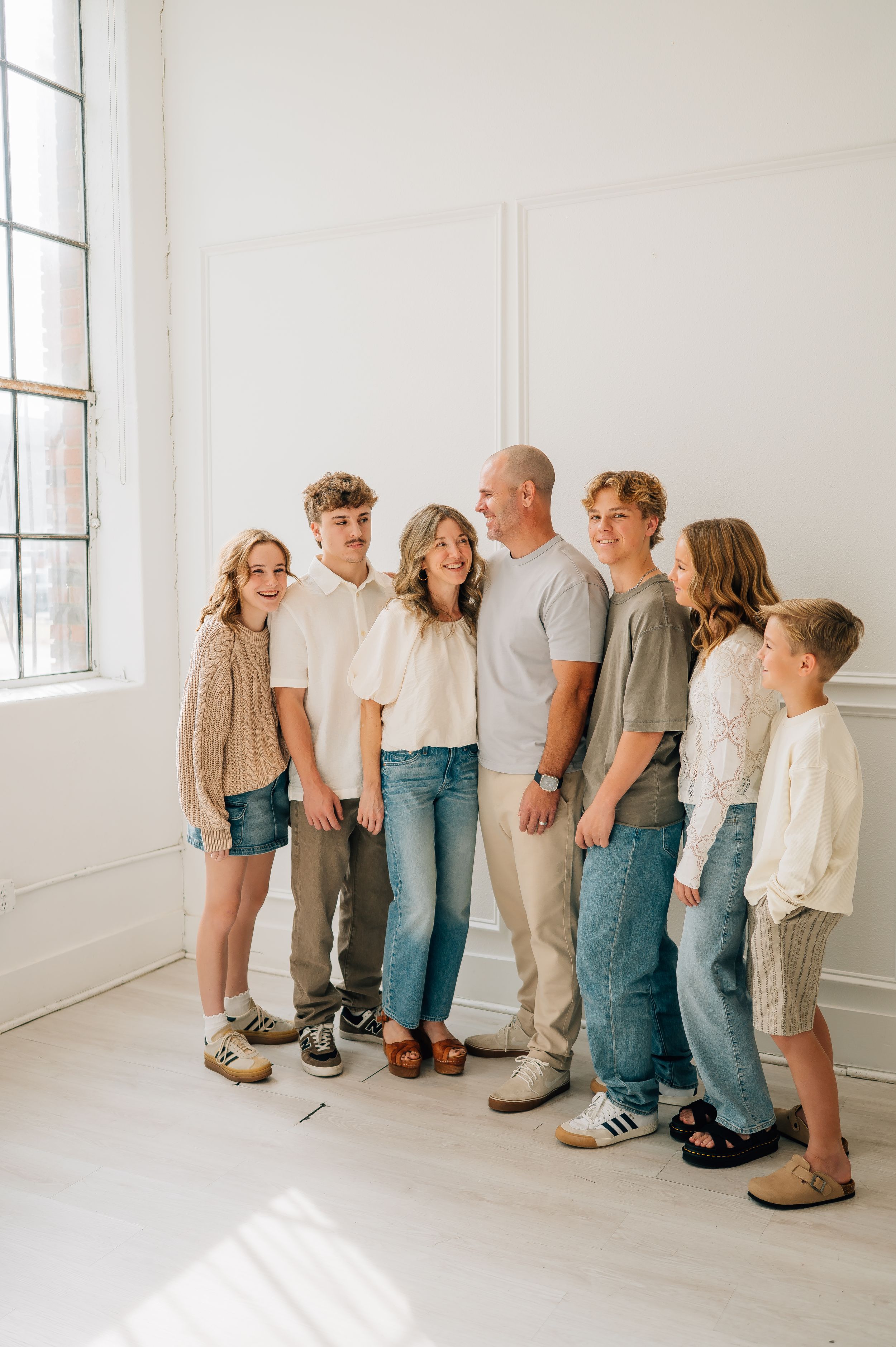 A family wearing coordinated casual outfits in neutral tones poses together in a bright white minimalist room.