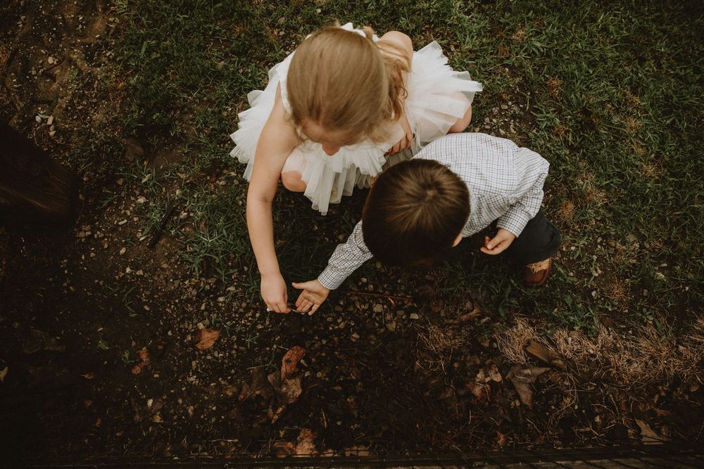 Flower girl and ring bearer crouch in the grass, curiously exploring the ground together during a wedding day moment.