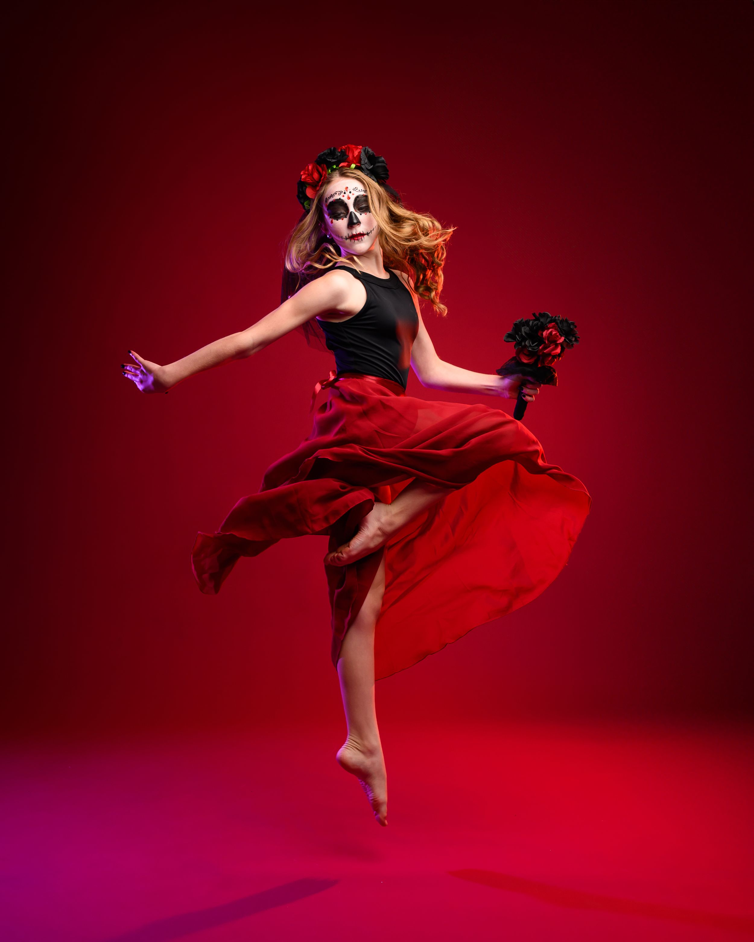 Dancer in Day of the Dead mask and flowing red dress performing graceful movements against crimson background.
