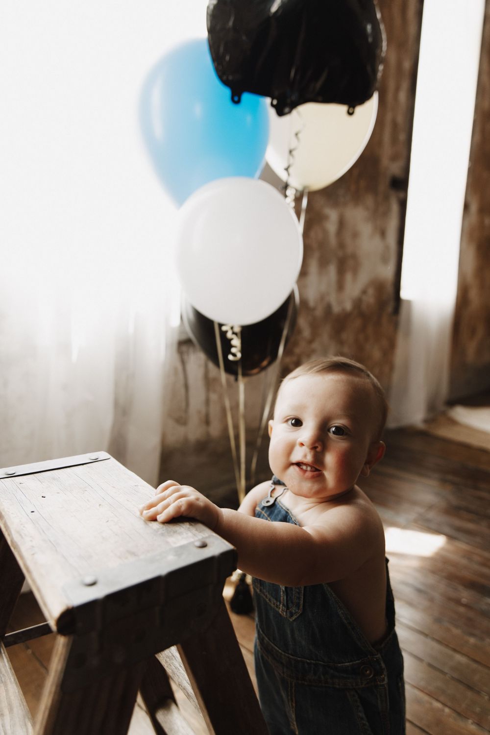 Baby in denim overalls sits near party balloons on hardwood floor during first birthday celebration.