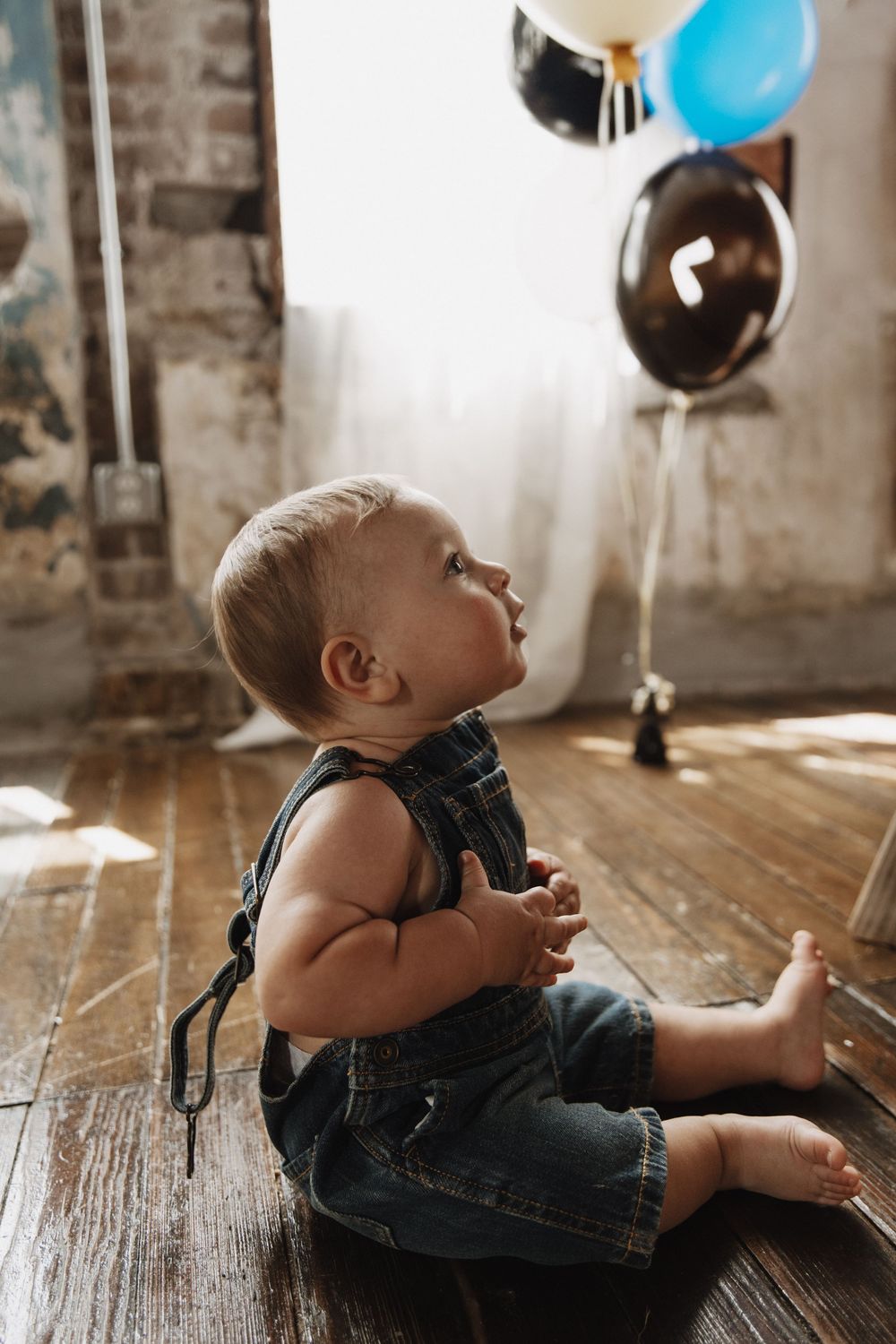 Baby in overalls plays with colorful balloons while lying on vintage hardwood floor in natural light.