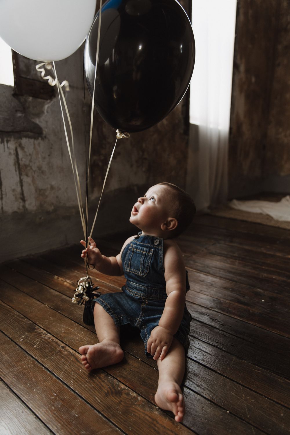 Baby in denim overalls lies on wooden floor holding white balloons in a moody rustic setting.