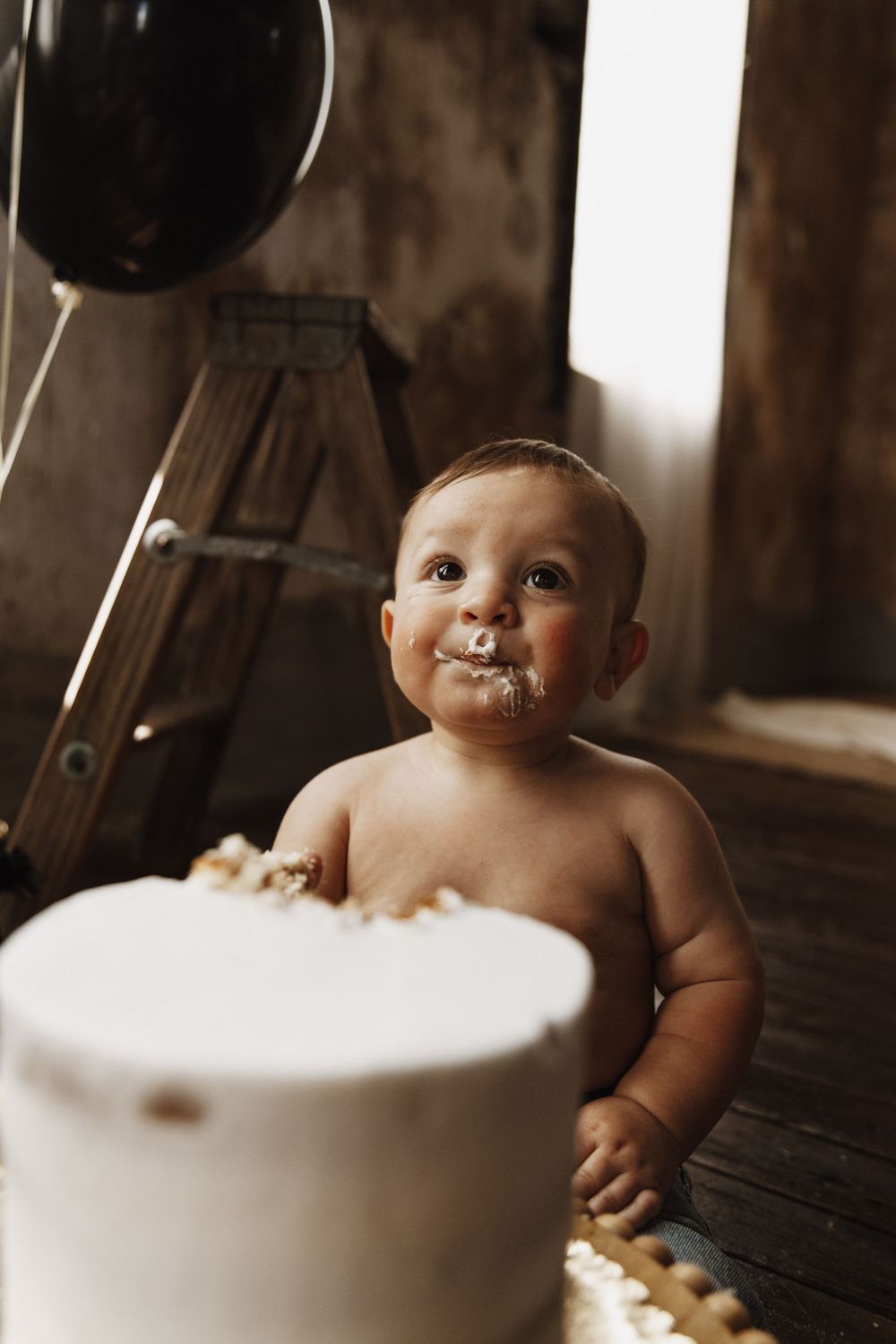 Baby sits at wooden table with white birthday cake and messy frosting on face.