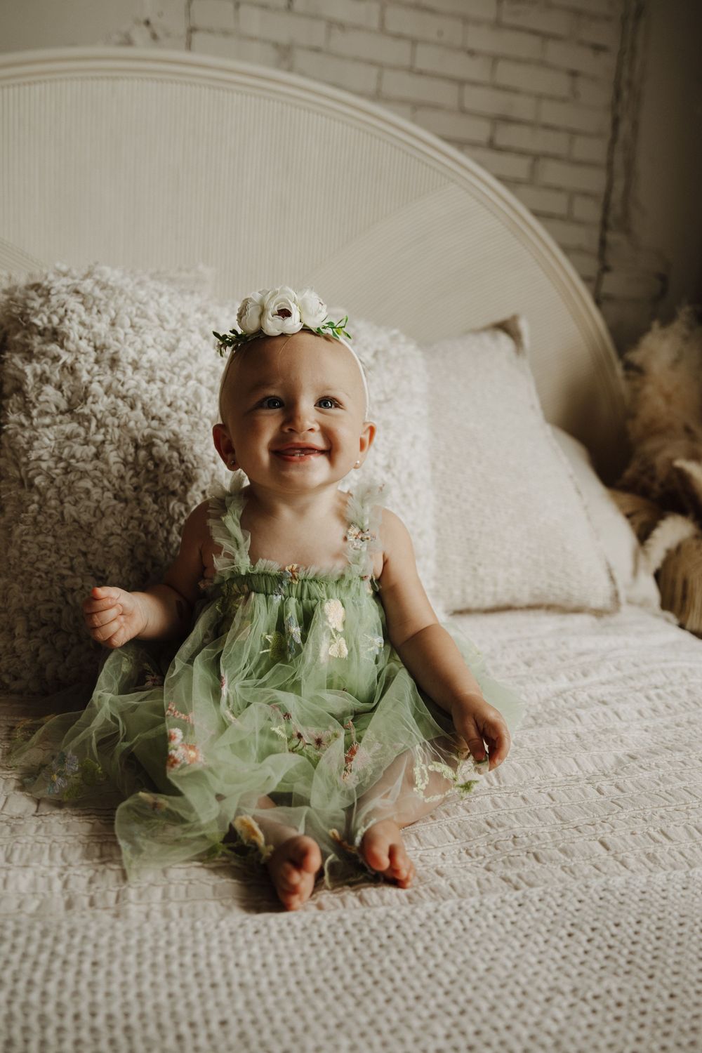 Small child wearing a mint green tulle dress sits and smiles on a white textured chair against a brick wall.