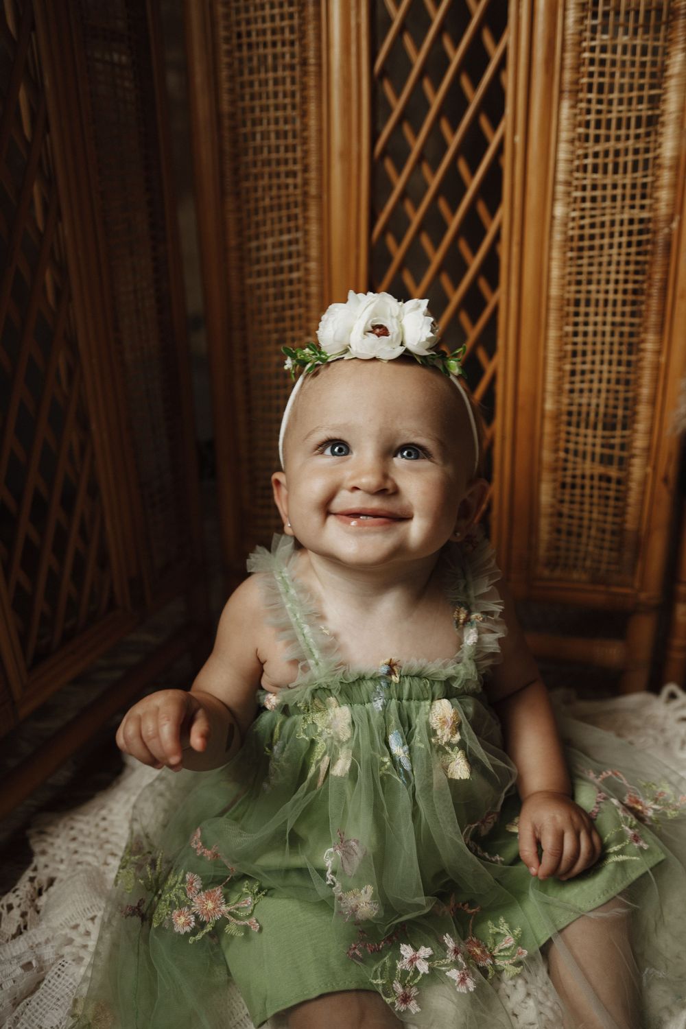 A baby in a sage green tulle dress with delicate floral details and a matching flower crown smiles while sitting.