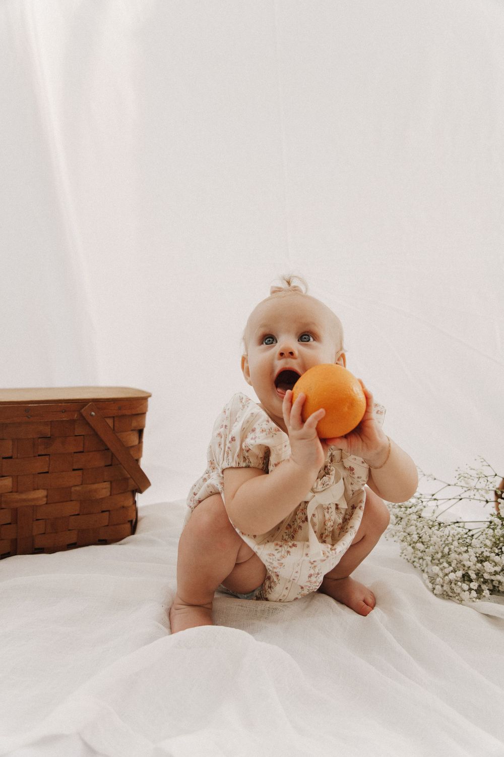 A baby in a lace dress reaches for and holds an orange fruit while sitting on white fabric near a brown wicker basket.