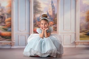 Child in light blue princess dress sits on floor in elegant room with painted scenery backdrop and white columns.