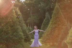 A girl in a fitted lace gown poses with arms outstretched between evergreen trees in the Hudson Valley,NY.