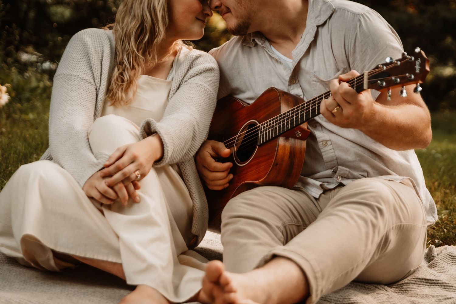 Romantic couple sits on grass while man plays acoustic guitar at sunset in casual attire.