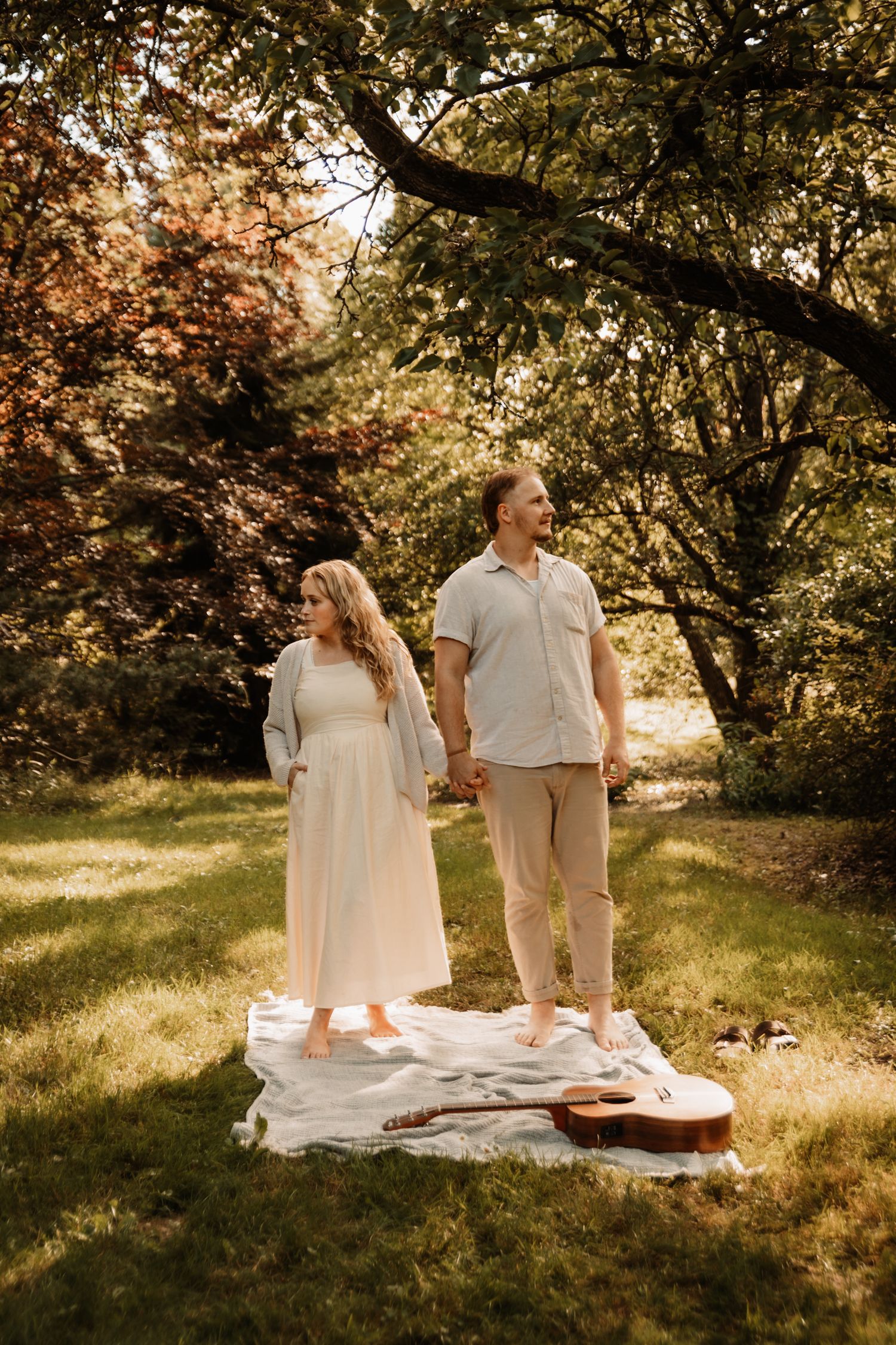 A couple stands on a picnic blanket in a sunlit park with a guitar lying nearby.